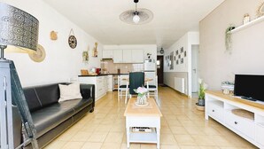 A modern living room with a sofa, coffee table, and TV stand. A kitchen is visible in the background with white cabinets and a dining area. - Renovated house and tastefully decorated in La Chaume (Les Sables-d´Olonne)