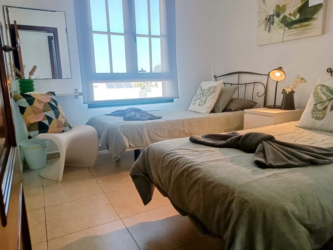 A bedroom with two beds, a chair, a bedside table with a lamp, and a window with blinds.
