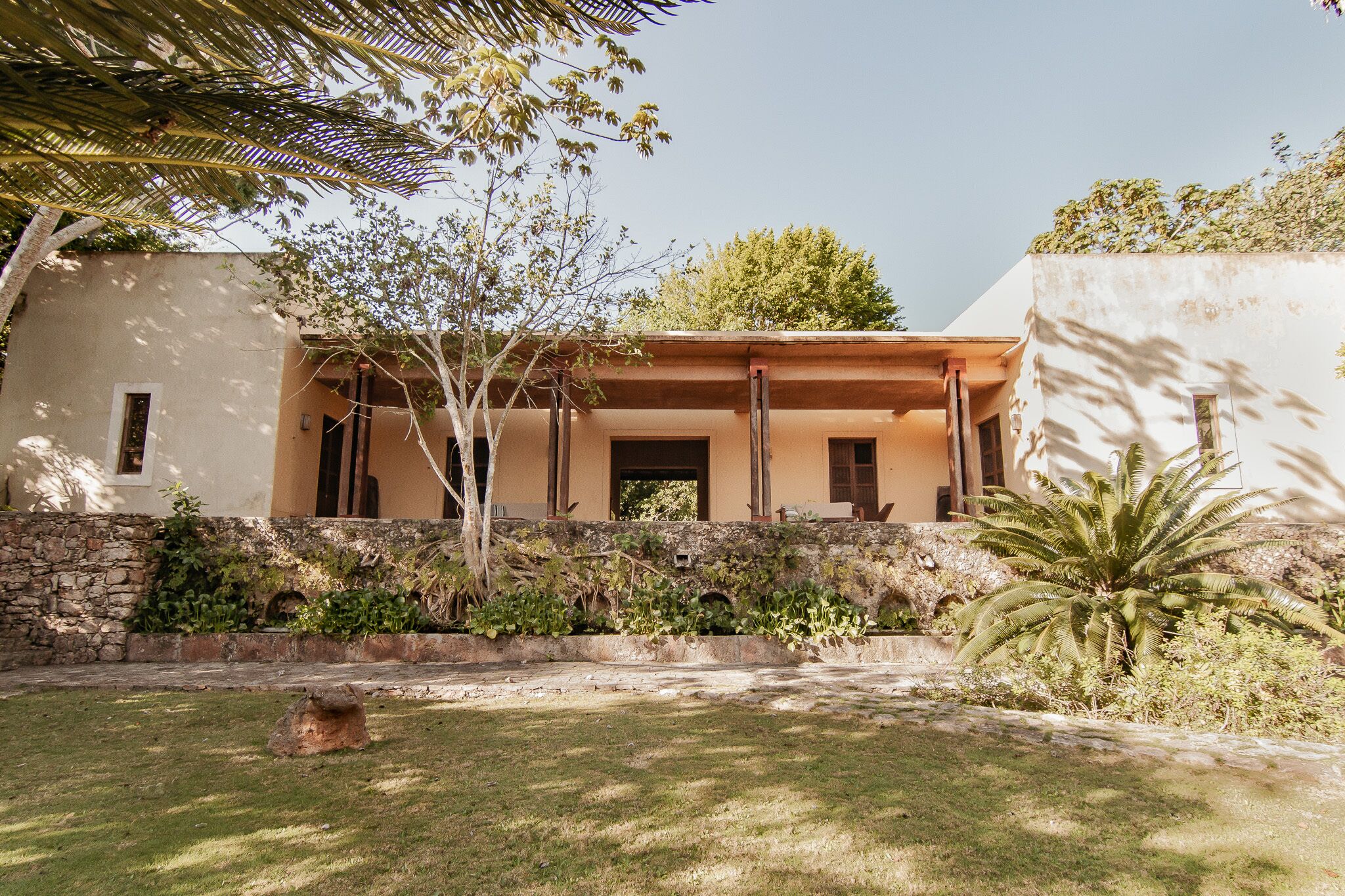 Un edificio con muro de piedra, un patio cubierto y una zona con césped y árboles.