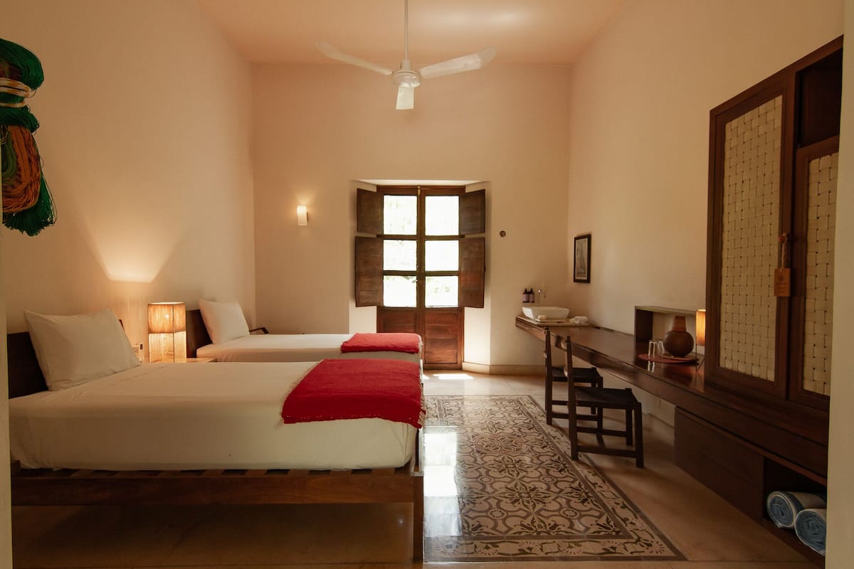 A hotel room with two beds, a desk, a chair, a ceiling fan, a window, and a patterned rug.
