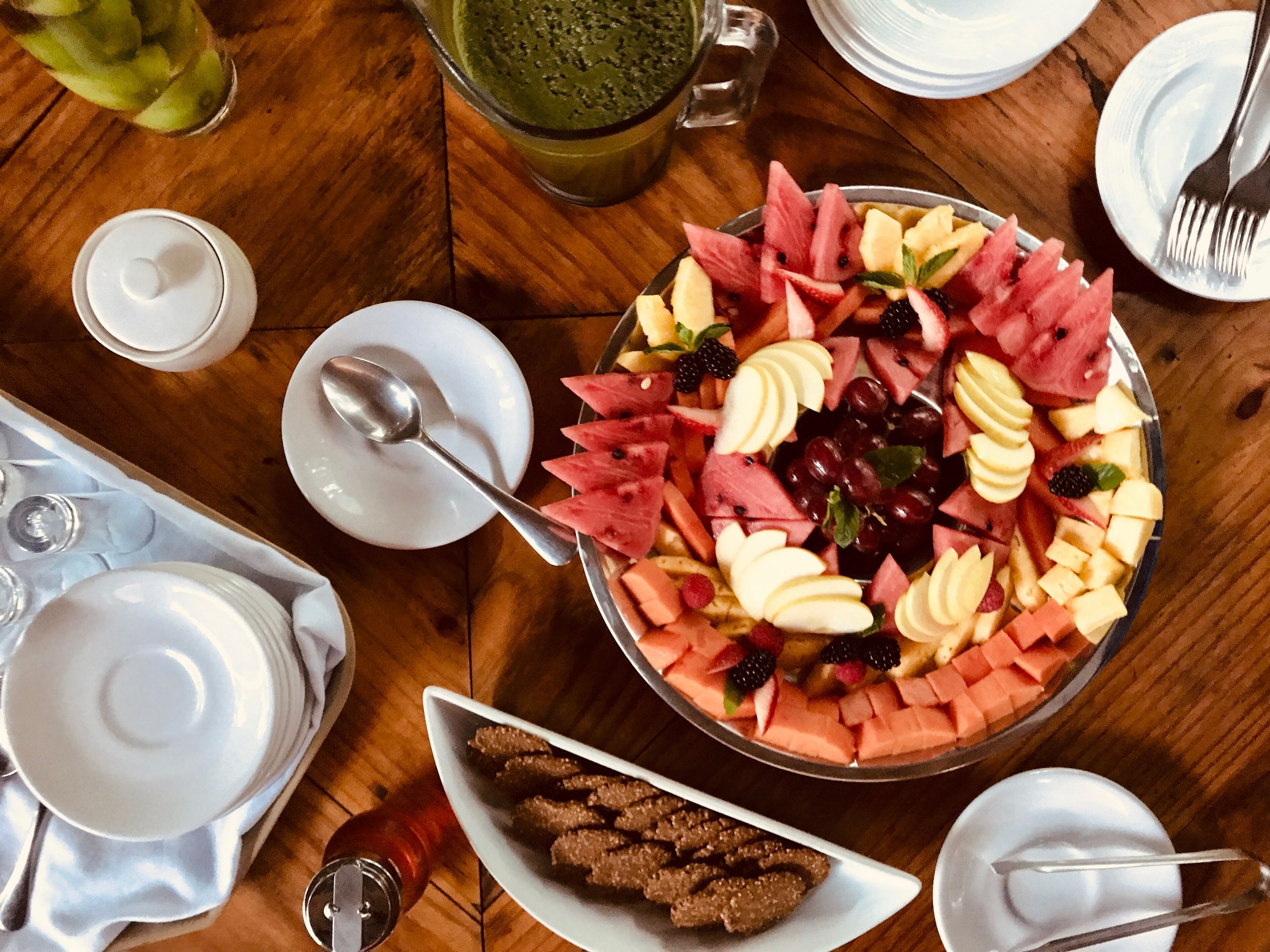Una mesa con una generosa bandeja de frutas, un vaso de jugo verde y diferentes platos y cubiertos.