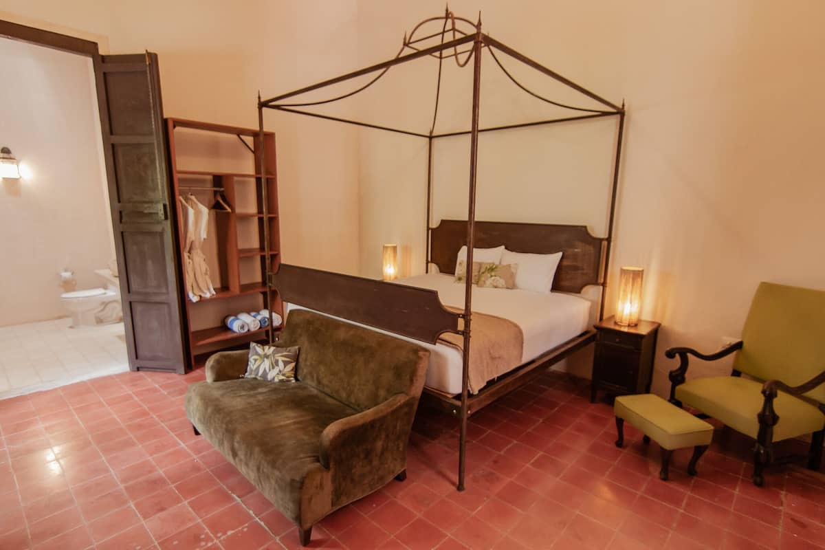 A four-poster canopy bed with a wooden headboard, a green armchair, a small bench, and a wooden wardrobe with a mirror.