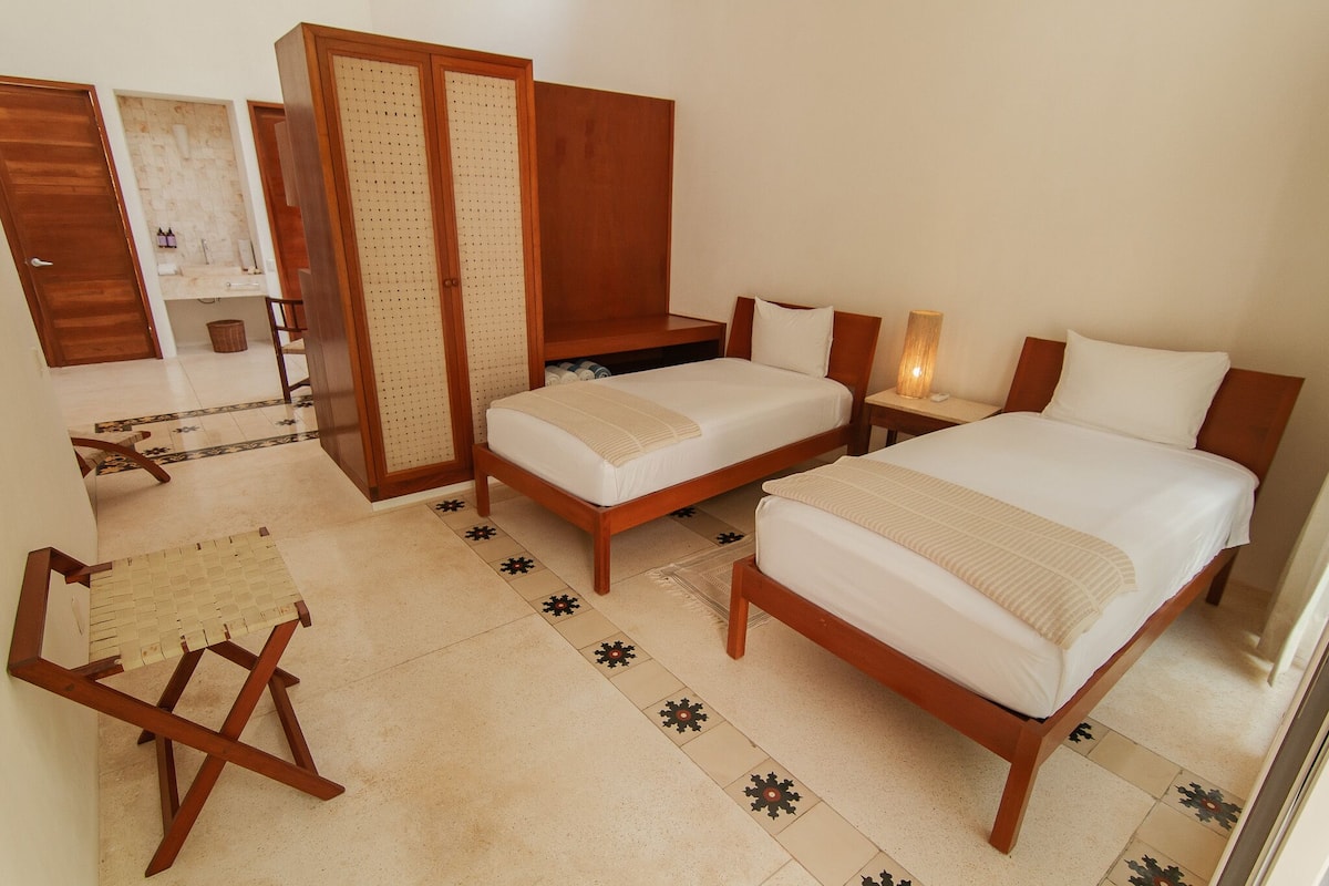 A hotel room with two beds, a wooden wardrobe, a small table with a lamp, and a woven chair.