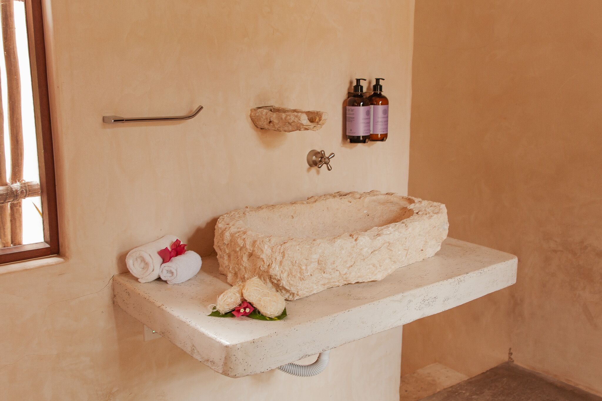 A bathroom with a stone sink, towels, toiletries, and a window.