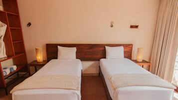 A hotel room with two single beds, wooden headboards, bedside lamps, a wooden shelf with towels, and a window with curtains.