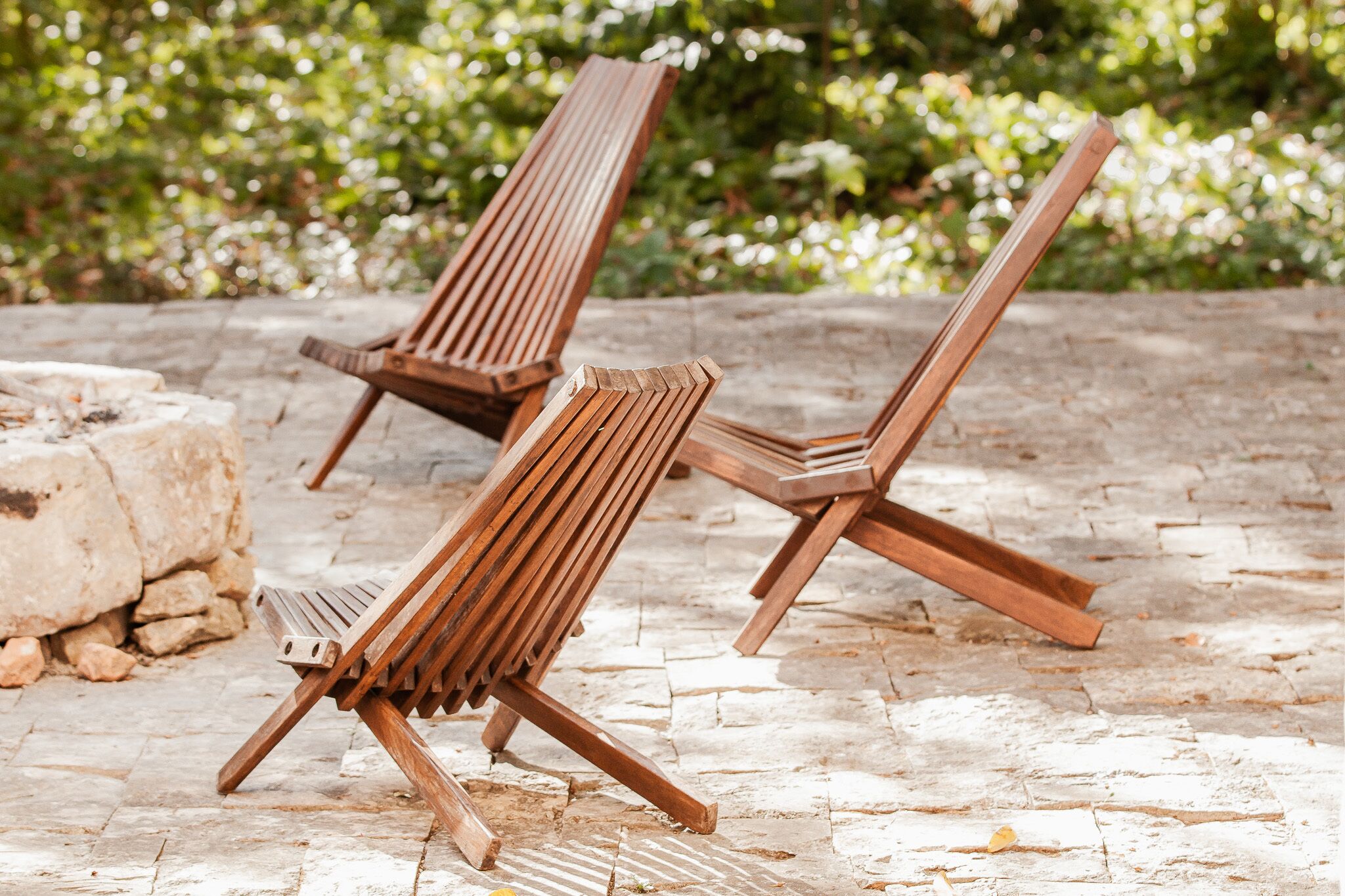 Zwei Holz-Loungesessel mit Metallgestellen stehen auf einer steingepflasterten Terrasse mit einer Steinmauer und Grünpflanzen im Hintergrund.