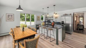 Une cuisine moderne dotée d’un îlot central, de tabourets de bar, de luminaires suspendus et d’un coin repas avec une table et des chaises en bois.
