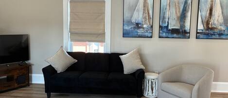 A living room with a black sofa, a beige armchair, a TV, and three sailboat paintings on the wall.