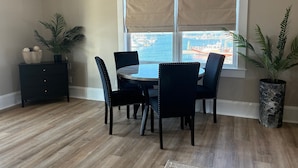 A dining area with a round table, four chairs, a dark wooden cabinet, and a view of the marina through a window with beige curtains. - The Captain's Quarters located in Cape Charles, VA offers breathtaking views. (Cape Charles)