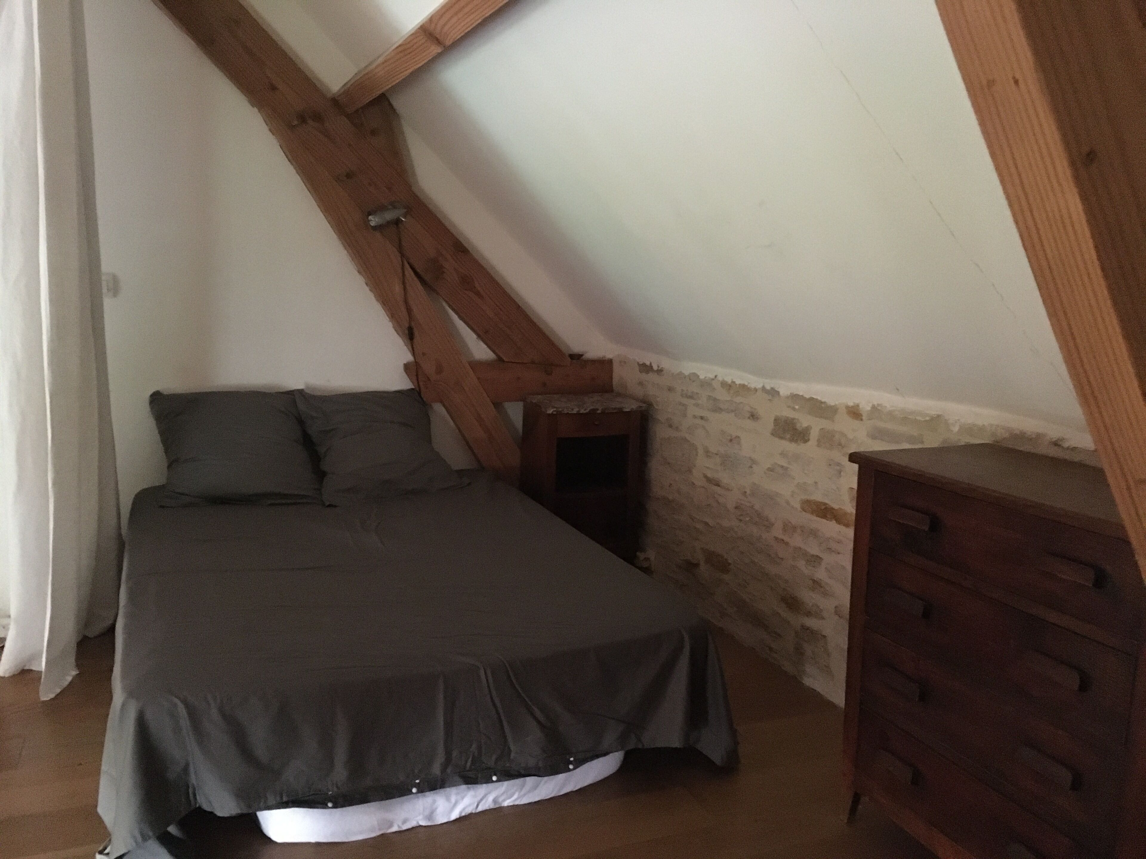 Ein Schlafzimmer mit schrägverdeckter Decke, einem Bett mit dunkler Bettdecke, einem hölzernen Schminktisch und einem kleinen Nachttisch.