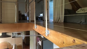 Une chambre de style « loft », avec un plancher en bois, un lit recouvert d’une couverture grise et une vue sur le salon.