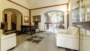 A spacious living area with a white sofa, a coffee table, and a fireplace. There is a staircase leading to another level, and a large window offering a view of the outdoors.