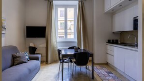 A compact living space with a sofa, dining table, chairs, a flat-screen TV, a kitchen area, and a window with curtains.