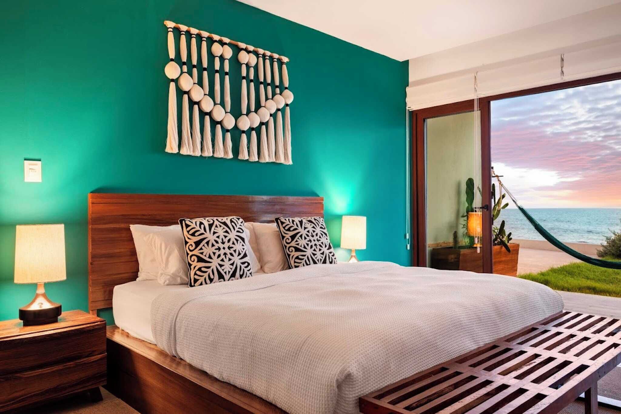 A bedroom with a bed, two bedside tables, a wall hanging, and a view of the ocean.