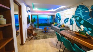 A modern living room with a view of the ocean, a dining area with a wooden table, and a large wall mural. - Malkita Sol | Oceanview Paradise + Concierge Service (Isla Mujeres)