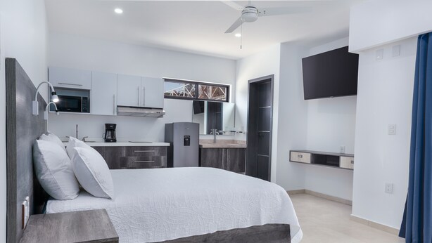 Un dormitorio moderno con una cama grande, un escritorio empotrado, un televisor de pantalla plana y una kitchenette al fondo.