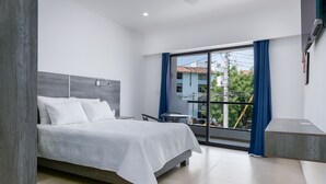 Un dormitorio moderno con una cama grande, una mesita pequeña y un balcón con vistas a edificios y árboles.