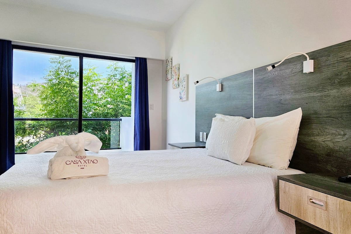 Une chambre d’hôtel avec un grand lit, une table de chevet, une lampe et une vue sur les arbres par la fenêtre.