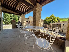 Un patio techado al aire libre con piso de piedra, sillas de metal, una mesa redonda y vistas a un edificio de piedra y vegetación.