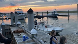 A wooden dock with people sitting, overlooking a marina with boats and a sunset in the background.