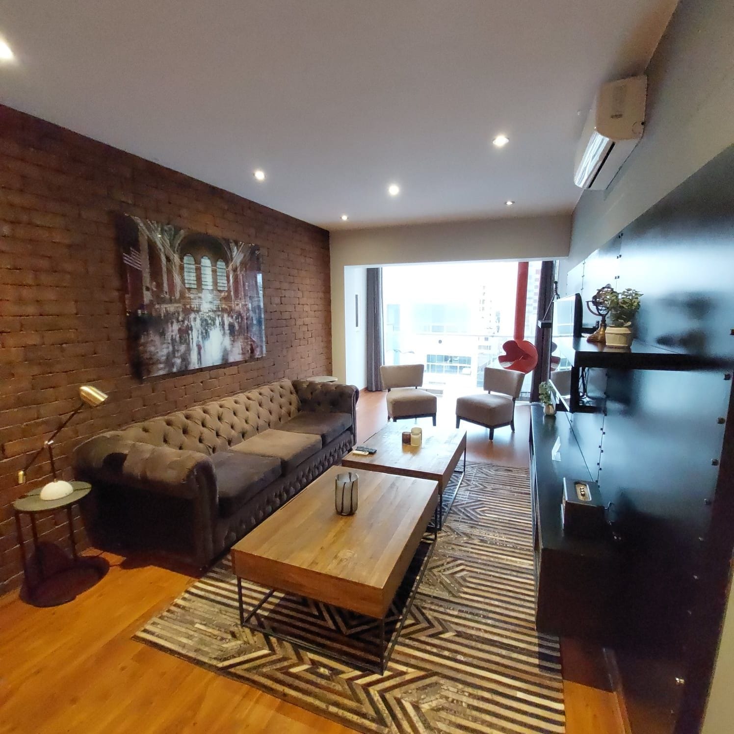 A modern living room with a sofa, coffee table, and chairs. There is a large window providing natural light and a view of the city.