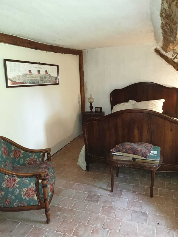 Une chambre à coucher avec un lit en bois, un fauteuil à motifs floraux, une petite table de chevet avec des livres et un tableau encadré accroché au mur.