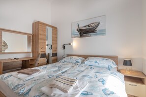 A neatly made bed with a blue and white nautical-themed duvet, a wooden nightstand with a lamp, a wooden desk with a mirror, and a wall-mounted painting of a boat. - Rückzugsort am Meer mit Pool, nur Wenige Minuten vom Strand Entfernt (Trget)