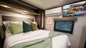 Una cama con cabecera, almohadas, una manta, un equipo de aire acondicionado, una ventana y un televisor montado en la pared.