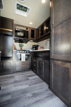 A compact kitchen with dark wood cabinets, a stainless steel range, a microwave, and a built-in oven. - Aileen Camping Space (Tampa)