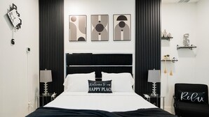 Ein modernes Hotelzimmer mit einem schwarzen Kopfteil, weißer Bettwäsche und einem Kissen mit der Aufschrift 'Welcome to HAPPY PLACE'. Über dem Bett befinden sich abstrakte Wandbilder, ein schwarzer Sessel mit einem Kissen mit der Aufschrift 'Relax' sowie ein kleines Regal mit Dekoration.