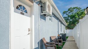Ein Haus mit weißer Tür, einer an der Außenwand angebrachten Klimaanlage, einer kleinen Terrasse mit zwei Korbstühlen und einer Fußmatte am Eingang.
