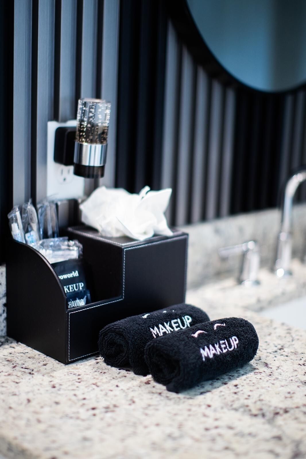 A bathroom with a black towel dispenser, a tissue box, and a pair of black slippers with 'MAKEUP' on them.