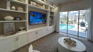 Een ruime woonkamer met een groot flatscreen televisietoestel, witte kasten en uitzicht op een zwembad door grote schuifdeuren.