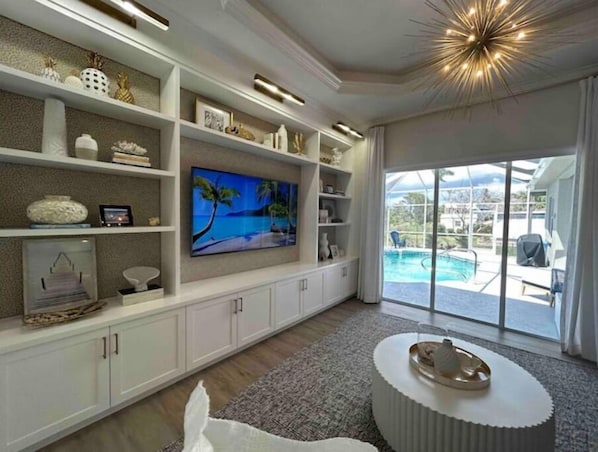 Ein geräumiges Wohnzimmer mit einem großen Flachbildfernseher, weißen Schränken und Blick auf einen Swimmingpool durch große Schiebeglastüren.