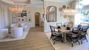 Een ruime woonkamer met een eethoek, met een grote houten tafel en rieten stoelen, een kroonluchter en uitzicht op de omgeving.