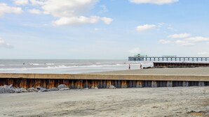 Una spiaggia con un molo che si protende nell'acqua, una barriera in cemento in primo piano e alcune persone che camminano sulla riva.