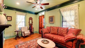 A room with a wooden floor, a red patterned sofa, a white armchair, a small table with a decorative bowl, and a ceiling fan. - Victorian Vacation Rental Apt in Downtown New Bern (New Bern)