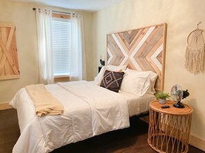 Een net opgemaakt bed met witte beddengoed en een beige plaid, een houten hoofdbord met een chevronpatroon, een klein rond nachtkastje met een ventilator, en een macramé wanddecoratie.