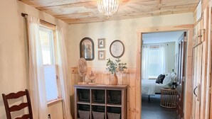 Una habitación con paneles de madera, una silla de madera, una consola con adornos y una cama al fondo.
