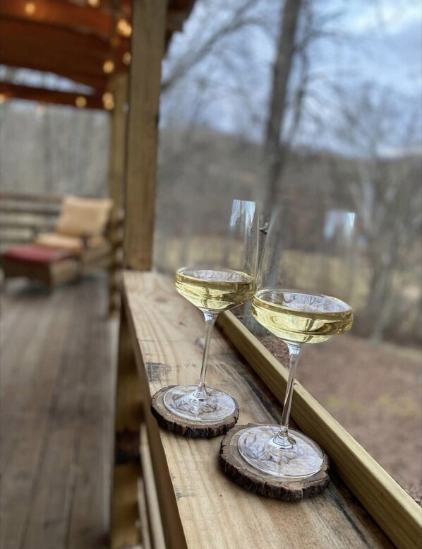 Dos copas de vino blanco sobre posavasos de madera, junto a una ventana con vista a una terraza de madera y sillas.