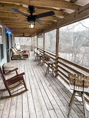Een houten terras met schommelstoelen en tafels, een plafondventilator en uitzicht op bomen door grote ramen.