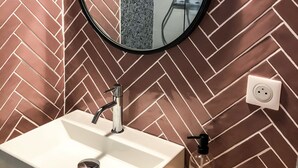 A bathroom with a herringbone-patterned wall, a round mirror, a white sink, and a handheld showerhead. - Studio in the historic center (Ajaccio)