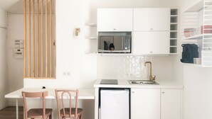A compact kitchen with white cabinetry, a microwave, a sink with a gold faucet, and a small refrigerator. There is a wooden partition and a chair in the dining area. - Studio in the historic center (Ajaccio)