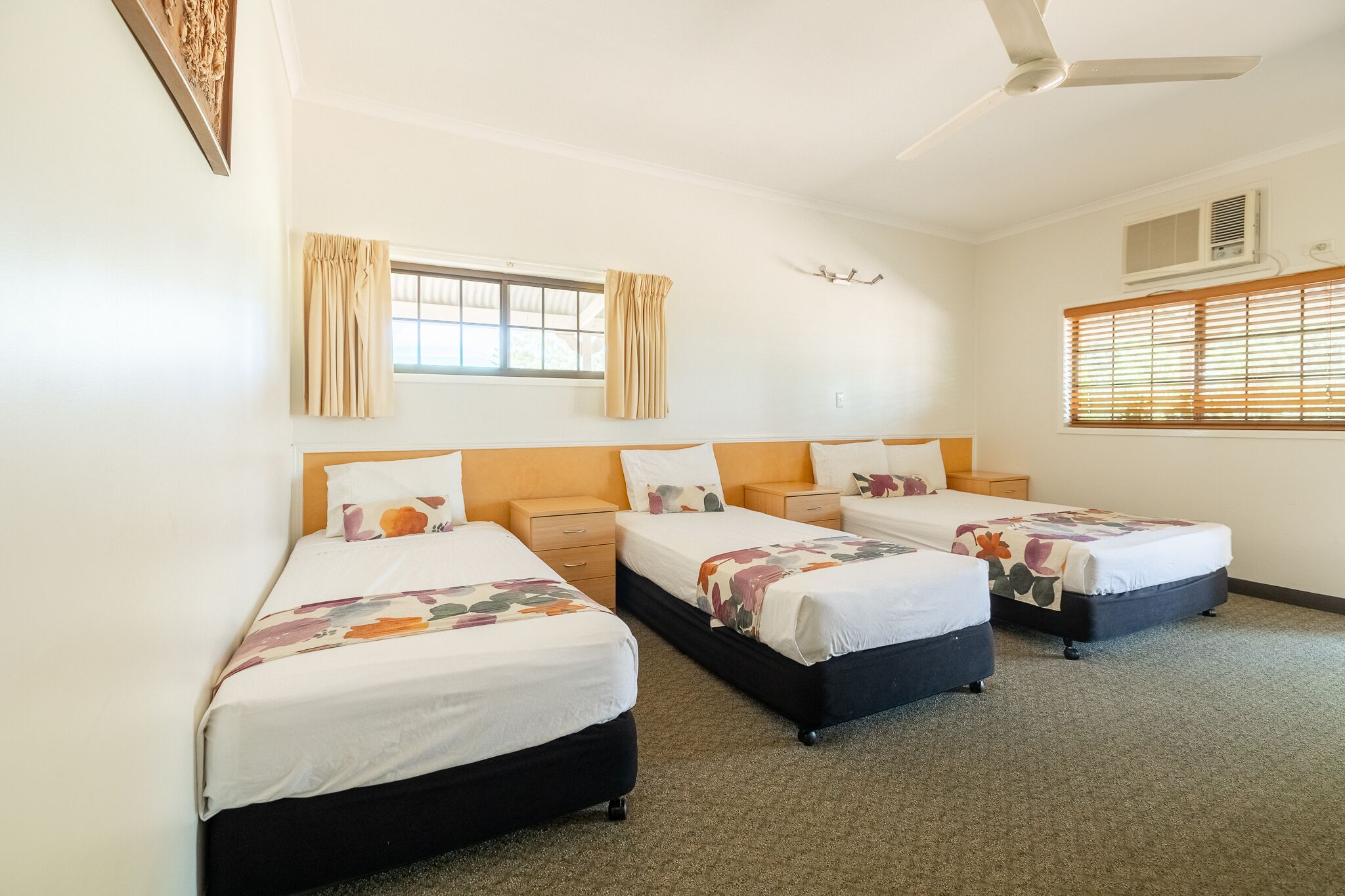 Une chambre d’hôtel avec trois lits simples, un ventilateur de plafond et une fenêtre avec des stores.