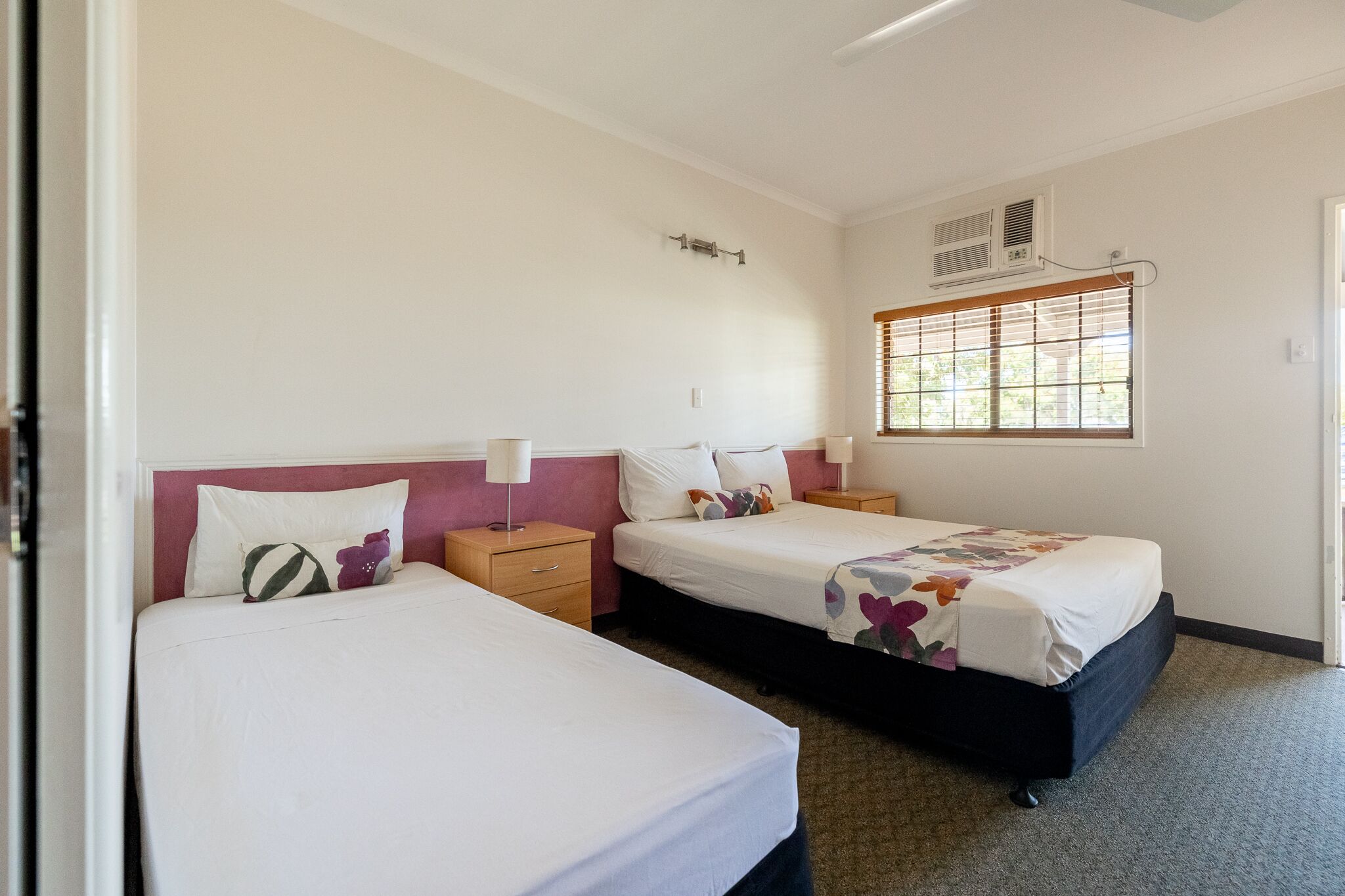 A hotel room with two beds, a window with blinds, an air conditioning unit, and a door.