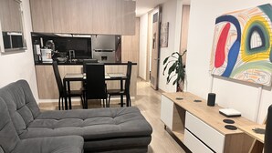 En moderne stue med en grå hjørnesofa, et træspisebord med stole, et køkken med køleskab og komfur, samt en væg med abstrakt kunst.