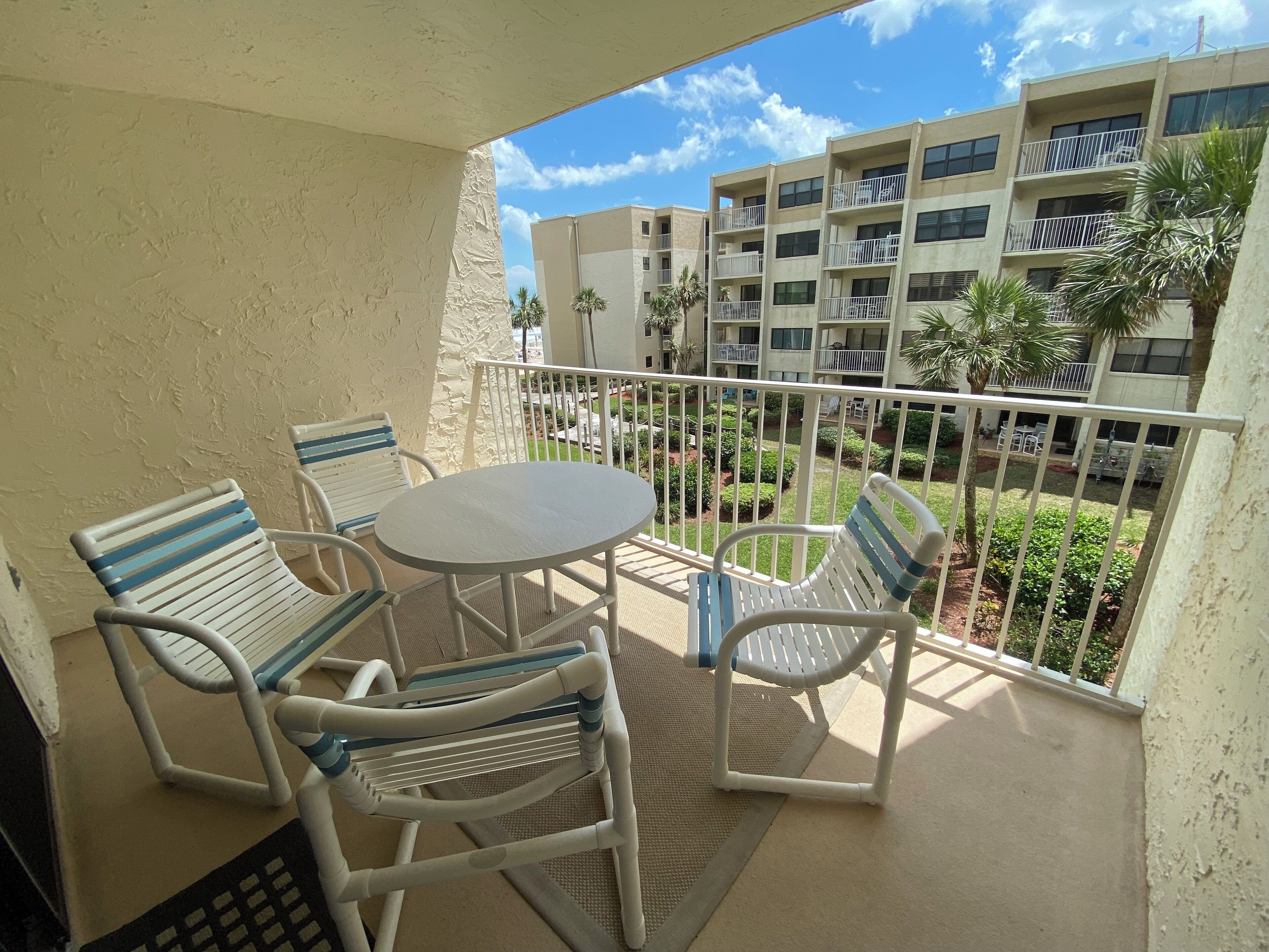 A balcony with white metal chairs and a table, overlooking a courtyard with greenery and a multi-story building in the background.