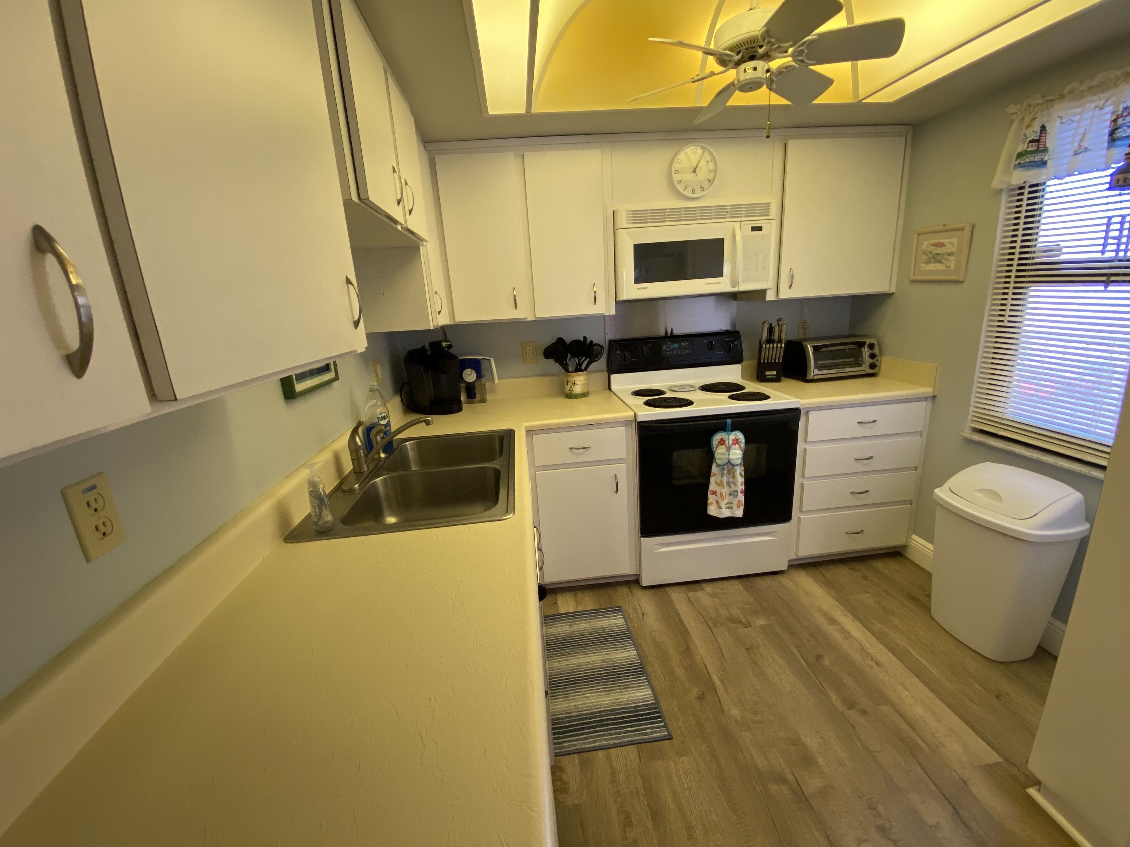 Una cocina funcional con armarios blancos, microondas, estufa, pileta y ventilador de techo.