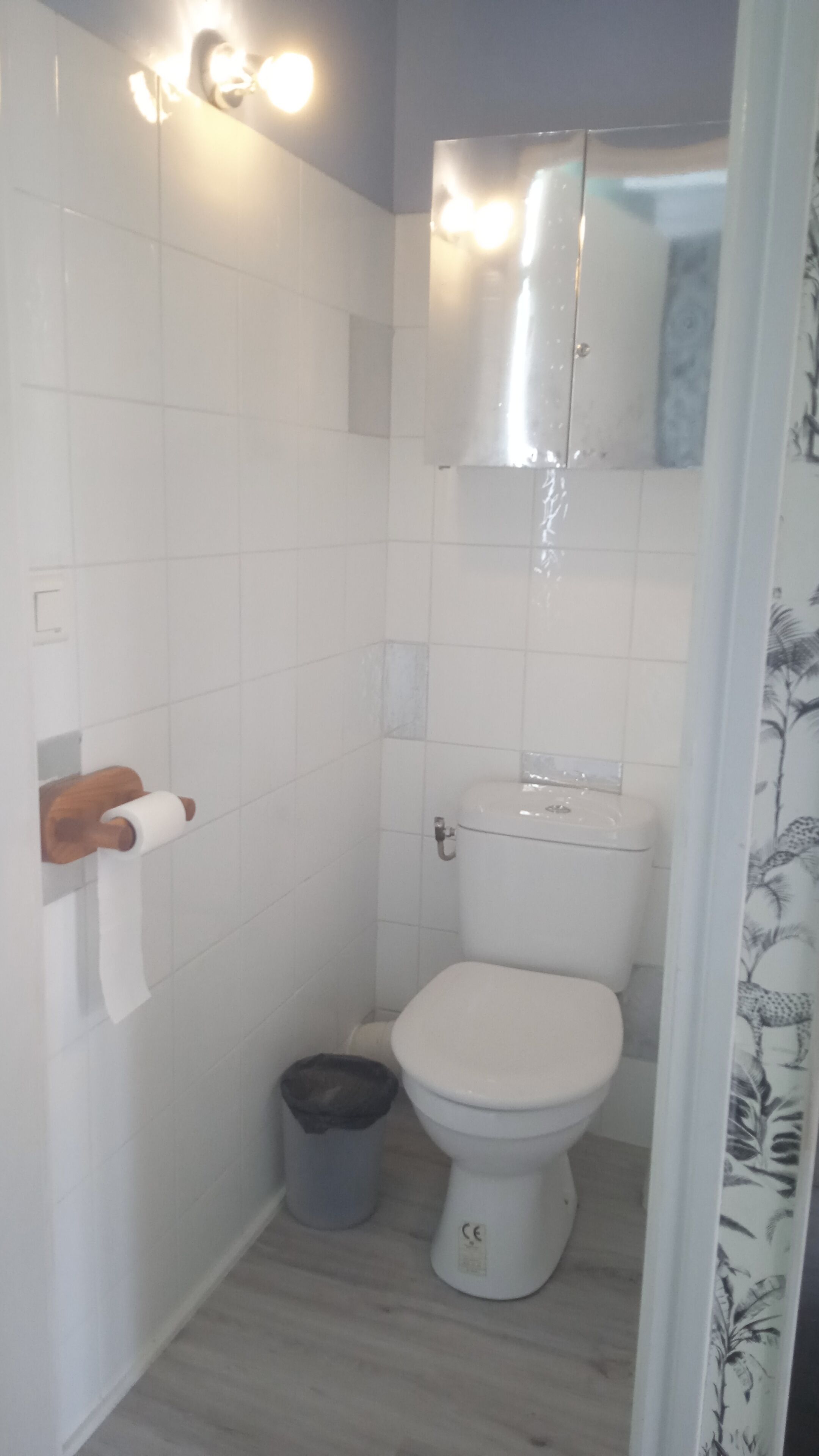Une petite salle de bain aux dimensions réduites, avec des carreaux blancs, des toilettes, une poubelle et un porte-rouleau de papier toilette en bois.