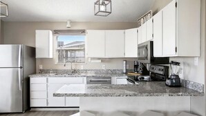 Une cuisine moderne avec des meubles blancs, un plan de travail en granit, un réfrigérateur, un four à micro-ondes, une cuisinière, un évier et une fenêtre donnant sur des immeubles.